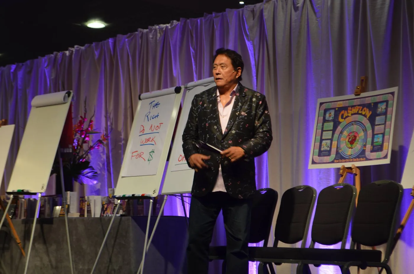 Trainer on a seminar stage with flip charts and a board-game poster, lit in purple