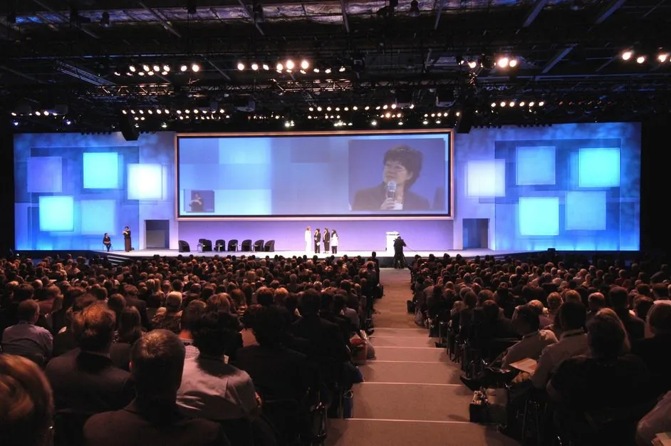 Seminar in a large hall: a panel on stage, a big central screen, lit set panels and a full auditorium