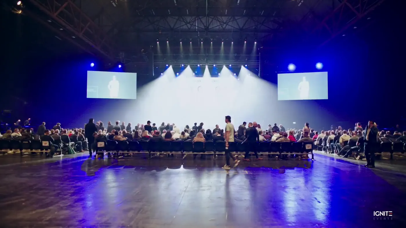 Large seated audience in front of a brightly lit stage with side screens