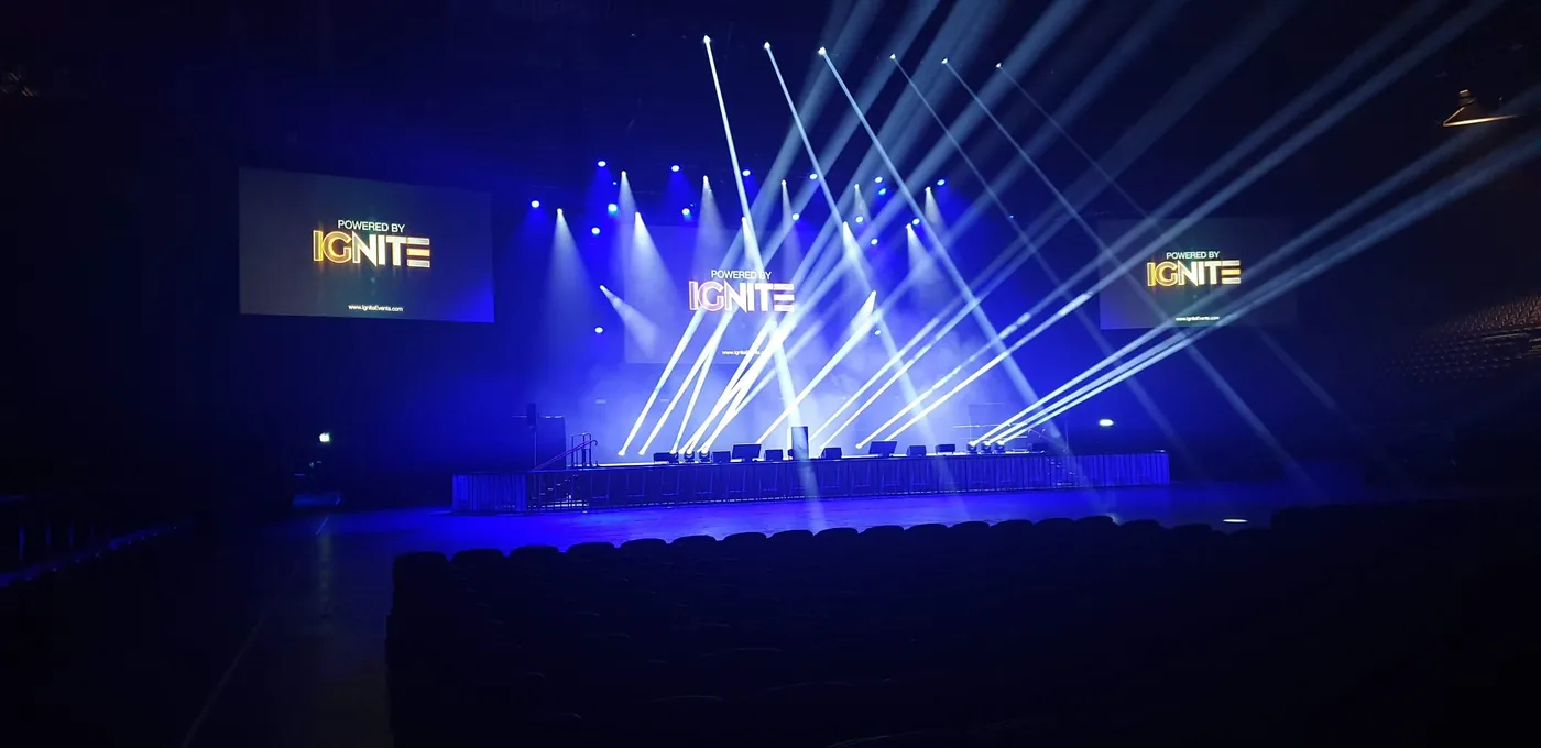 Moving-head beams crossing over an arena stage with 'Powered by IGNITE' on the screens