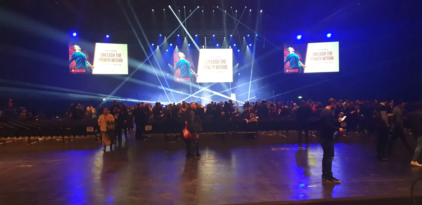 Arena audience on the floor between sessions, with beam lighting and three large screens above the stage