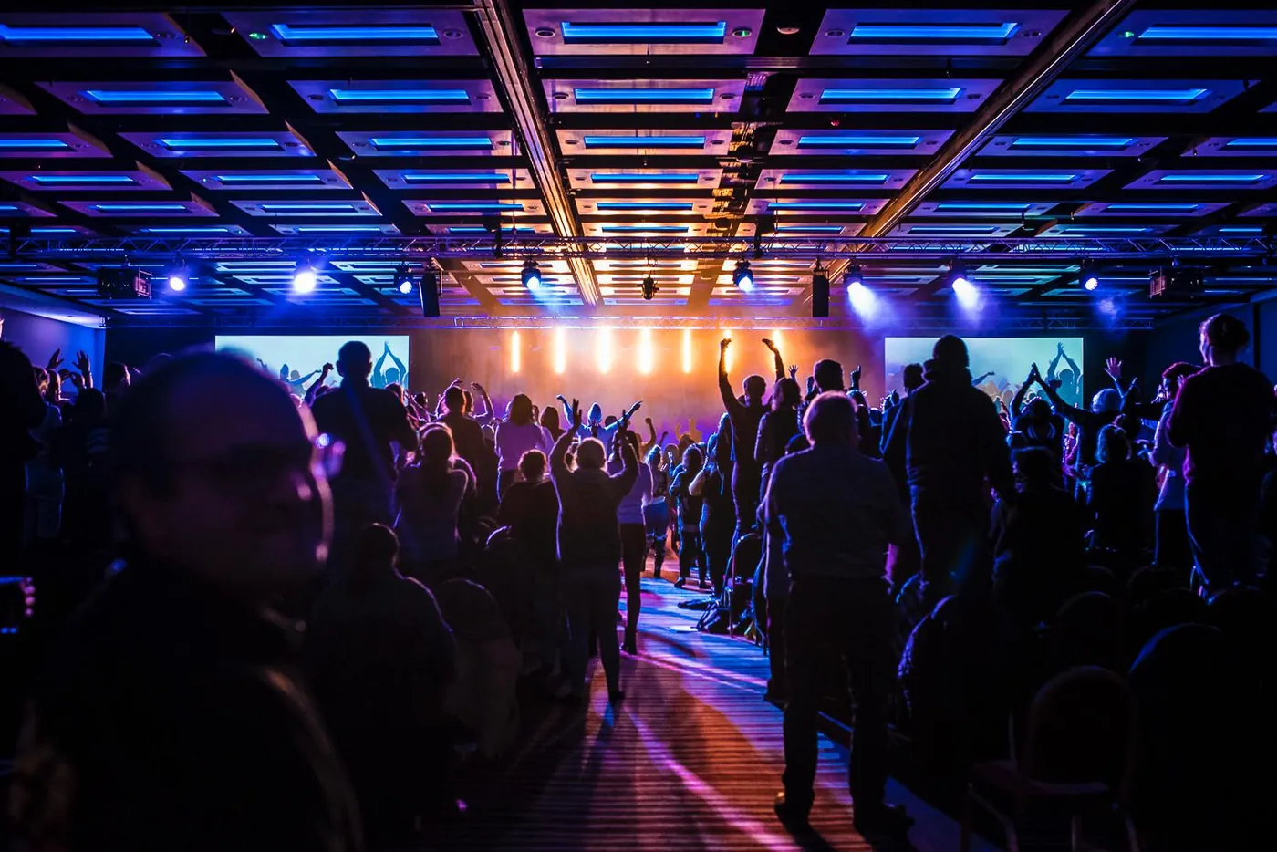 Audience on its feet with arms raised towards a lit stage and side screens at the ILEC Conference Centre, London