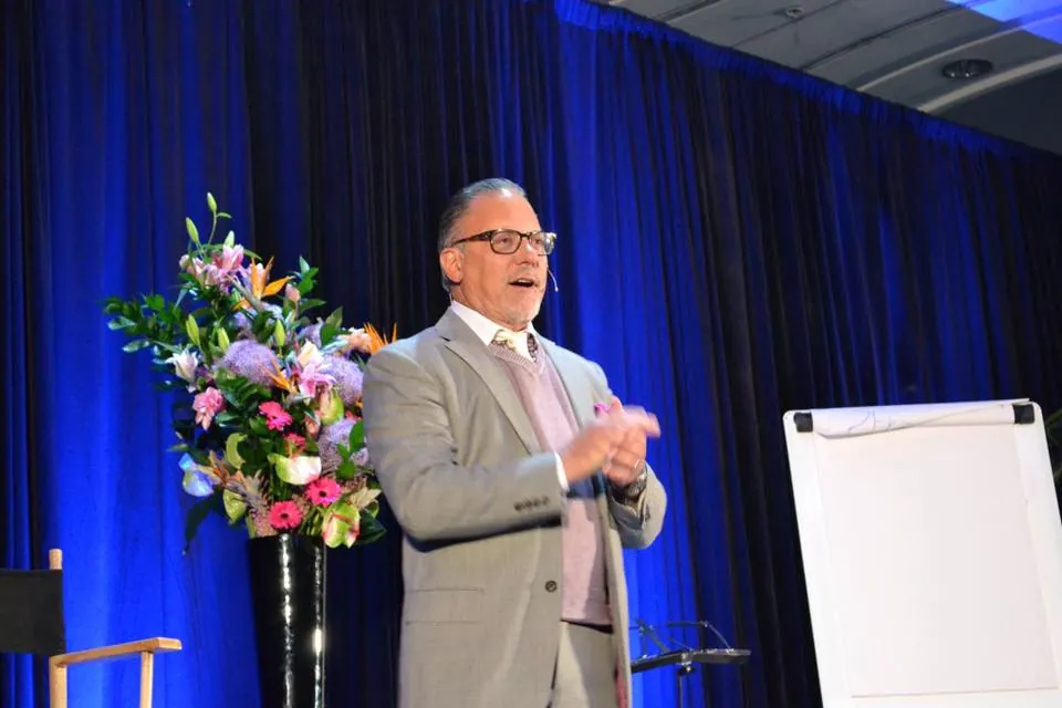 Speaker on stage in front of a blue curtain, with a flip chart and floral display