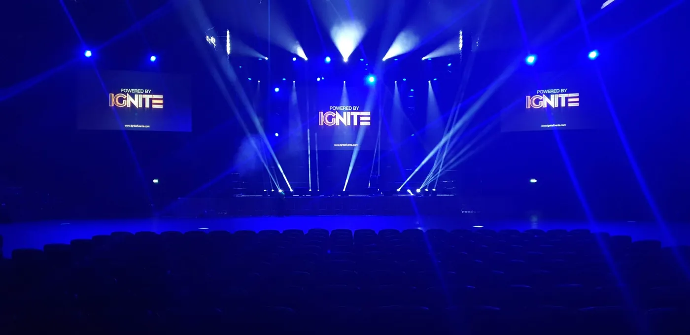 Arena stage in blue light with beam fixtures and 'Powered by IGNITE' on three screens