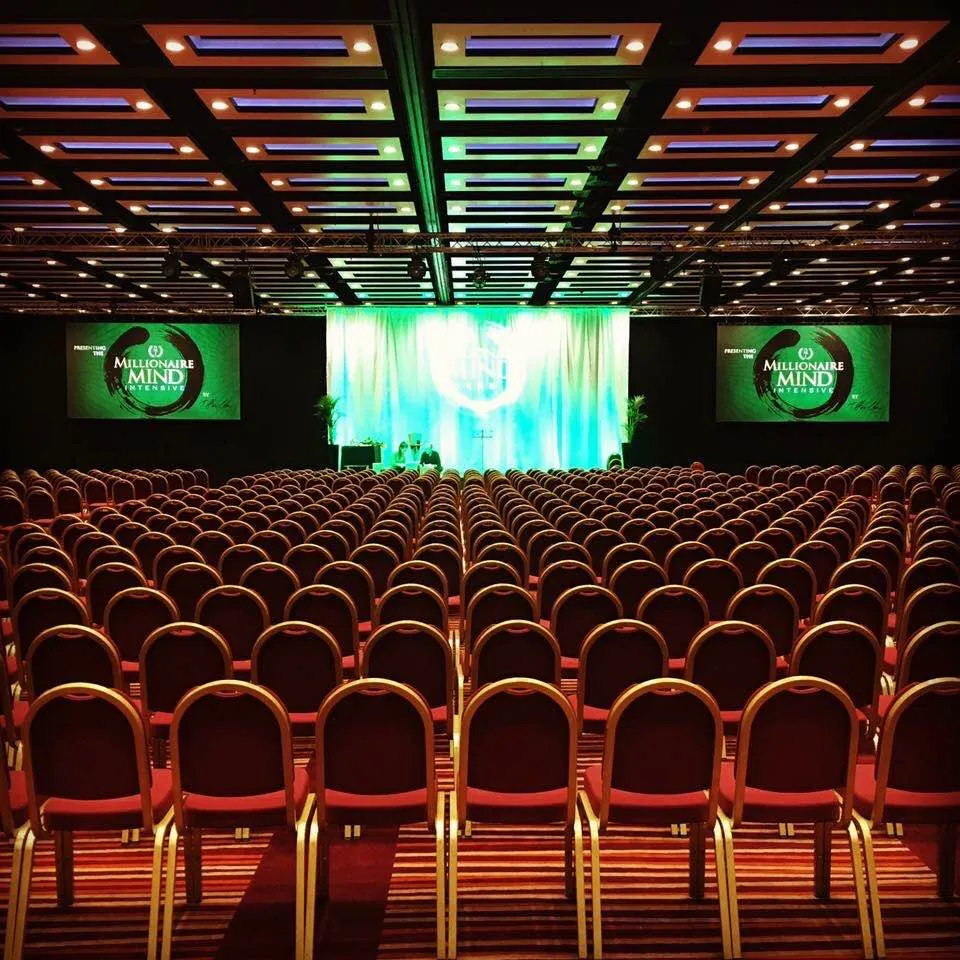 Main hall at the ILEC Conference Centre set theatre-style for the Millionaire Mind Intensive, with a green-lit stage drape and screens either side